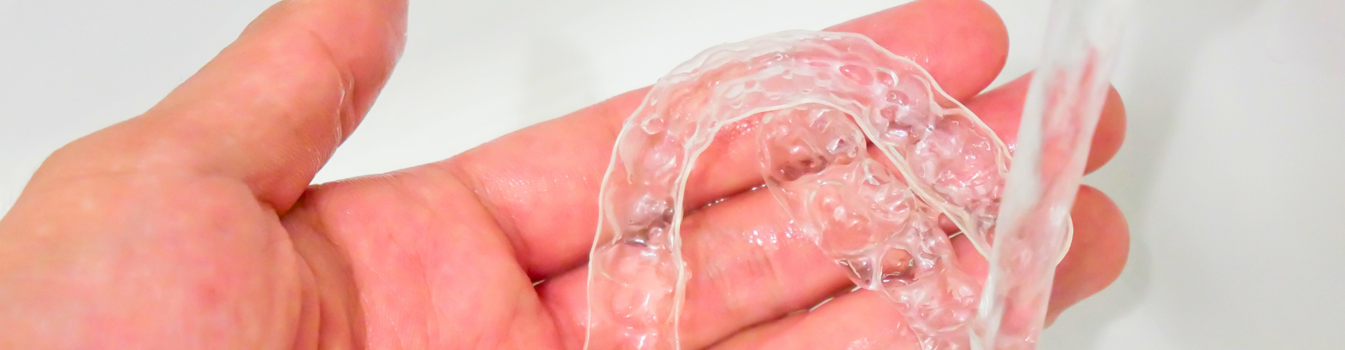 Hand holding clear aligner under water faucet.